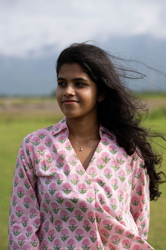 A woman with long dark hair stands outdoors in a grassy field wearing a pink button-up shirt with a green and pink floral block print pattern.