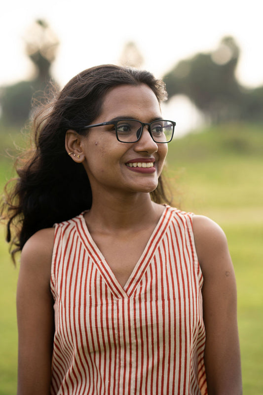 A woman with dark hair and glasses wearing a red and white vertically striped sleeveless top, smiling outdoors against a blurred green background.
