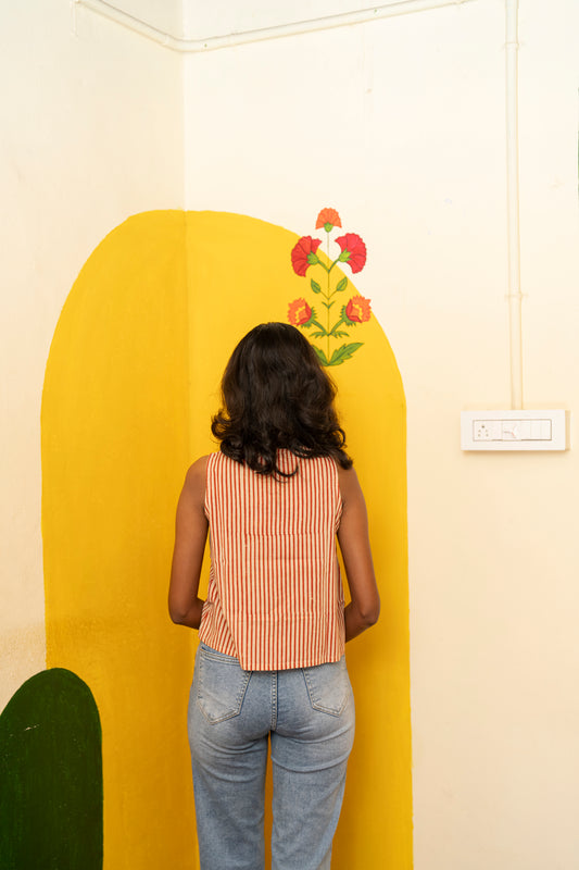 A woman viewed from behind stands in front of a bright yellow arched wall panel with a painted flower motif, wearing a sleeveless red-and-white striped top and light blue jeans.