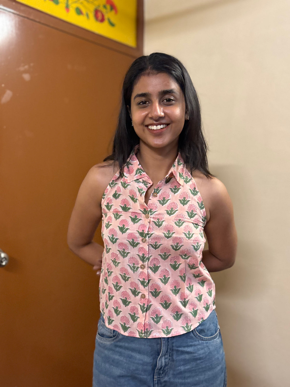 Woman wearing a pink floral top and blue jeans standing against a beige wall.