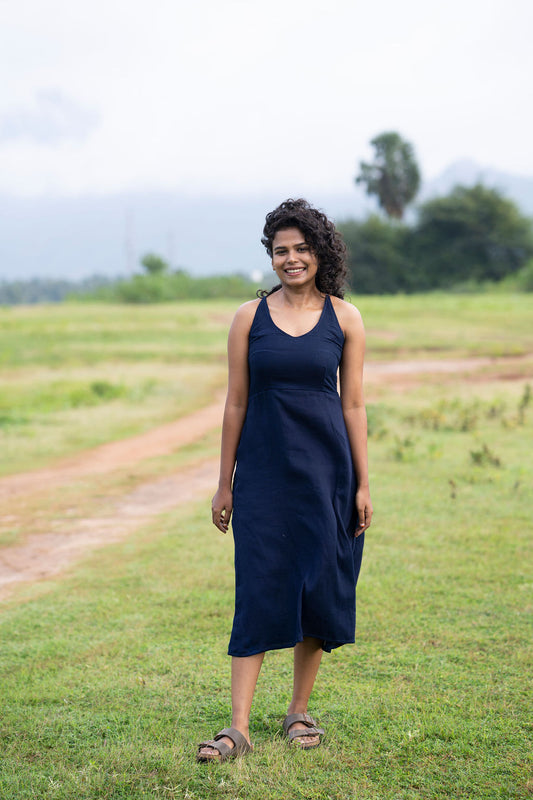 A woman wearing a sleeveless navy blue midi dress stands on a grassy field with a dirt path and distant trees under a cloudy sky.