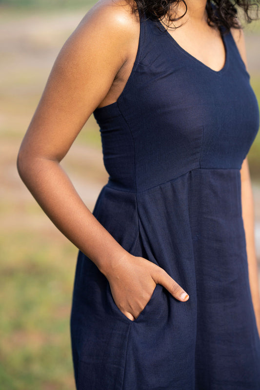 A close-up of a woman wearing a sleeveless navy blue dress with side pockets, standing outdoors with a blurred natural background.