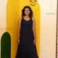 A woman stands barefoot in a room corner wearing a sleeveless dark navy midi dress. The background features a painted yellow arch on the wall, a green arch on the left, and a cream-colored wall with floral patterns.