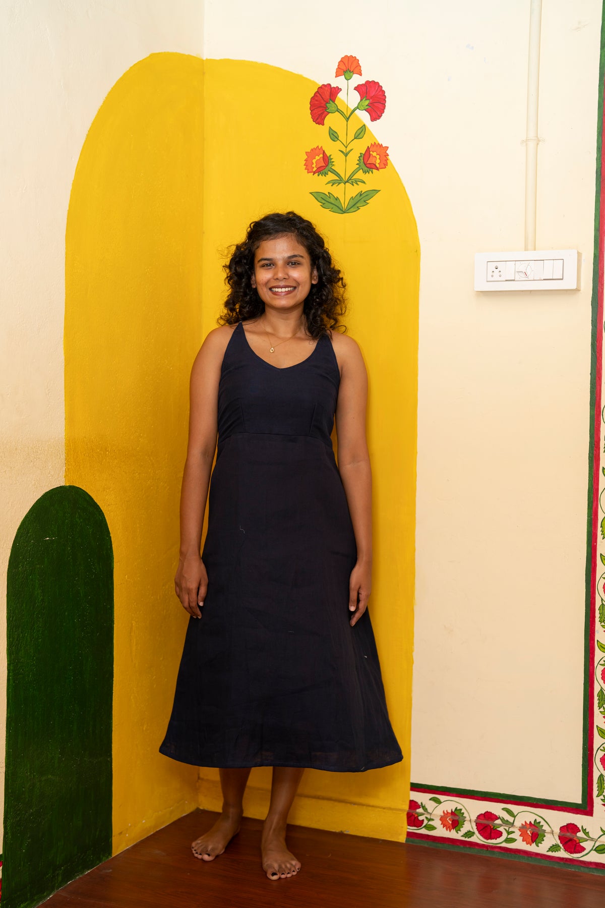 A woman stands barefoot in a room corner wearing a sleeveless dark navy midi dress. The background features a painted yellow arch on the wall, a green arch on the left, and a cream-colored wall with floral patterns.