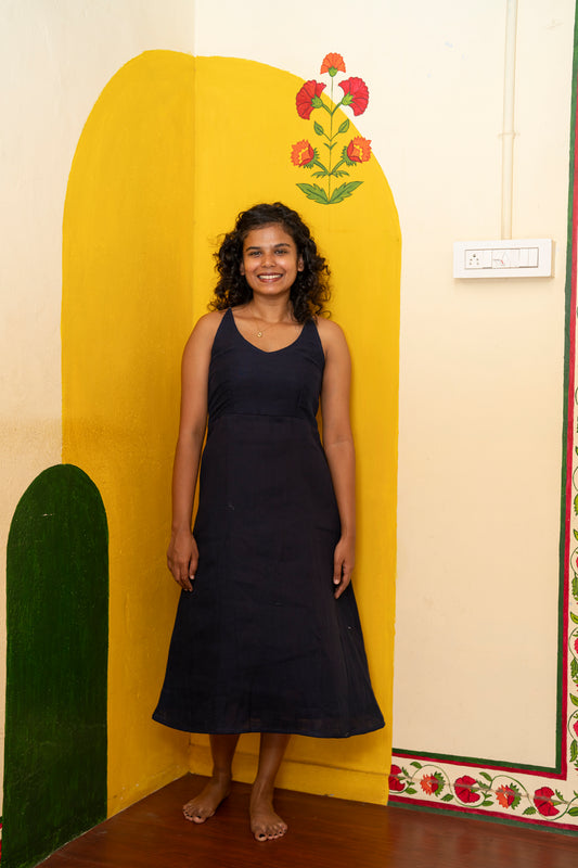 A woman stands barefoot in a room corner wearing a sleeveless dark navy midi dress. The background features a painted yellow arch on the wall, a green arch on the left, and a cream-colored wall with floral patterns.