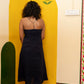 A woman with curly dark hair stands with her back to the camera wearing a strapless black midi dress, positioned in front of a bright yellow painted archway on a cream-colored wall.