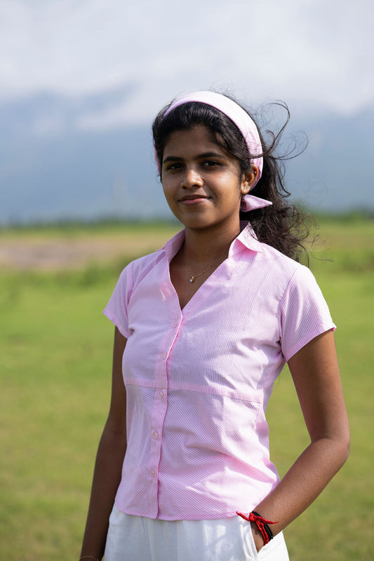 A young girl standing outdoors in a grassy field, wearing a light pink short-sleeve button-up shirt, a matching pink headband, and white pants.