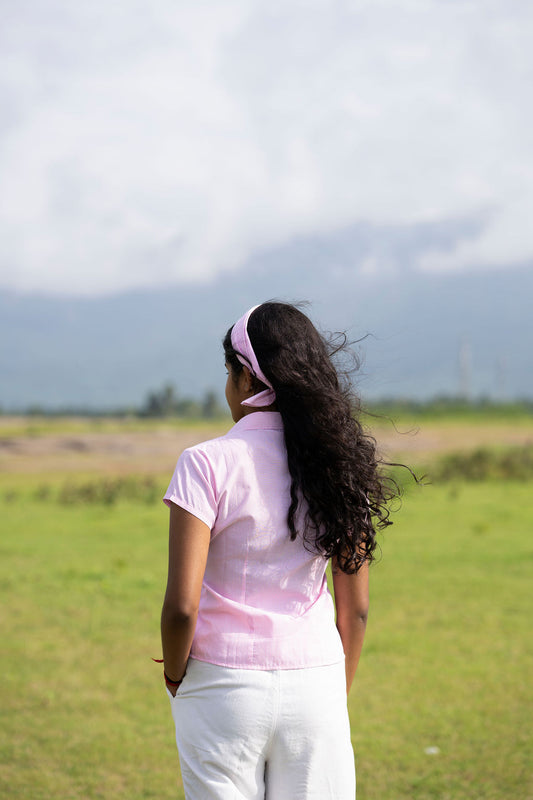 A woman seen from behind wearing a light pink short-sleeve collared shirt and white pants, standing in a grassy field with mountains and a cloudy sky in the background.