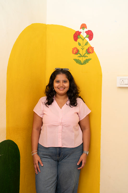 A woman with dark curly hair wearing a light pink short-sleeve button-up shirt and blue jeans, standing in front of a yellow arched wall with painted flowers.