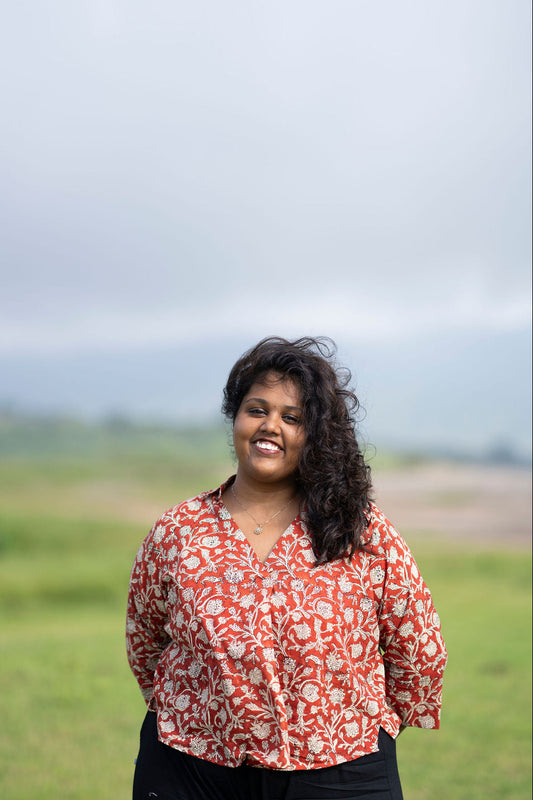 A woman with curly dark hair stands outdoors in a grassy field under a cloudy sky, wearing a red and white patterned blouse and black pants.