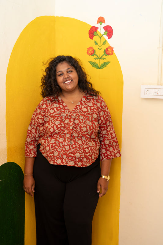 A woman with curly black hair stands smiling in front of a yellow arched backdrop with a floral design. She is wearing a red and beige patterned button-up shirt with three-quarter sleeves and black pants.
