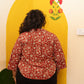 A person viewed from behind wearing a red and beige floral print shirt and black pants, standing in front of a large yellow arch-shaped wall panel decorated with a floral motif.