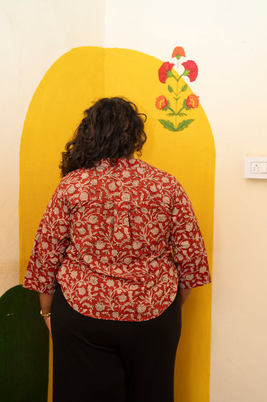 A person viewed from behind wearing a red and beige floral print shirt and black pants, standing in front of a large yellow arch-shaped wall panel decorated with a floral motif.