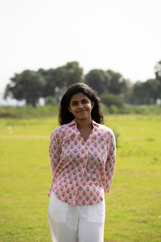 A woman standing outdoors in a grassy field wearing a pink patterned long-sleeve button-up shirt and white pants.