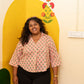 A woman smiling while wearing a pink and green patterned short-sleeve button-up shirt with black pants, standing in front of a yellow arched wall panel with a floral design.