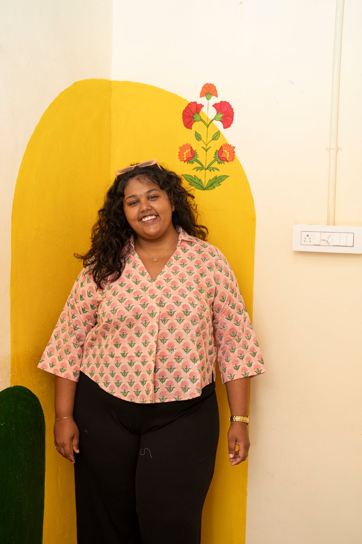 A woman smiling while wearing a pink and green patterned short-sleeve button-up shirt with black pants, standing in front of a yellow arched wall panel with a floral design.