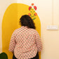A person viewed from behind wearing a light pink patterned shirt and black pants, standing in front of a yellow arched wall panel with a floral design.