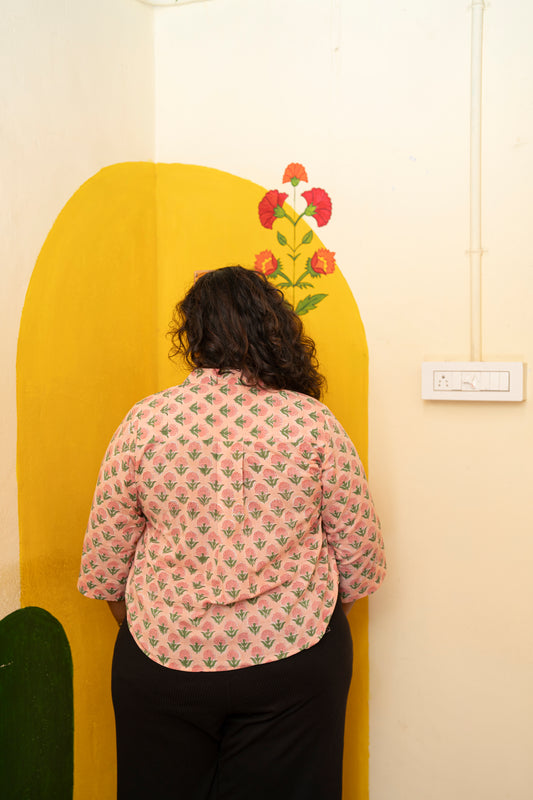 A person viewed from behind wearing a light pink patterned shirt and black pants, standing in front of a yellow arched wall panel with a floral design.