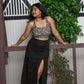 Woman wearing a black bralette top and black skirt standing against a decorative wall with plants.