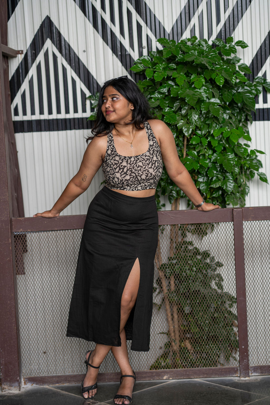 Woman wearing a black bralette top and black skirt standing against a decorative wall with plants.