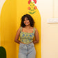 A woman with curly dark hair stands smiling in front of a bright yellow arched wall decoration. She is wearing a sleeveless green and blue patterned crop top and light-wash blue jeans.