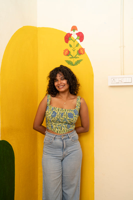 A woman with curly dark hair stands smiling in front of a bright yellow arched wall decoration. She is wearing a sleeveless green and blue patterned crop top and light-wash blue jeans.