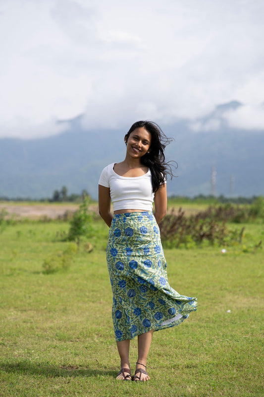 A woman stands in a grassy field with mountains and cloudy skies in the background, wearing a white cropped tank top and a blue and green floral patterned maxi skirt.