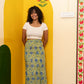 A woman stands barefoot in a room corner wearing a white short-sleeve crop top and a green and blue floral print midi skirt, posing in front of a yellow painted archway.