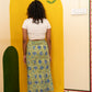 A woman stands with her back to the camera wearing a white cropped t-shirt and a green and blue floral patterned midi skirt, posing in front of a yellow arched wall panel with a painted flower motif.