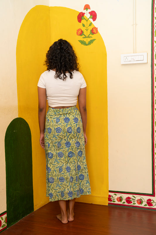 A woman stands with her back to the camera wearing a white cropped t-shirt and a green and blue floral patterned midi skirt, posing in front of a yellow arched wall panel with a painted flower motif.