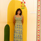 A woman stands barefoot in a room corner wearing a sleeveless yellow and blue floral print maxi dress, posing in front of a painted yellow archway.