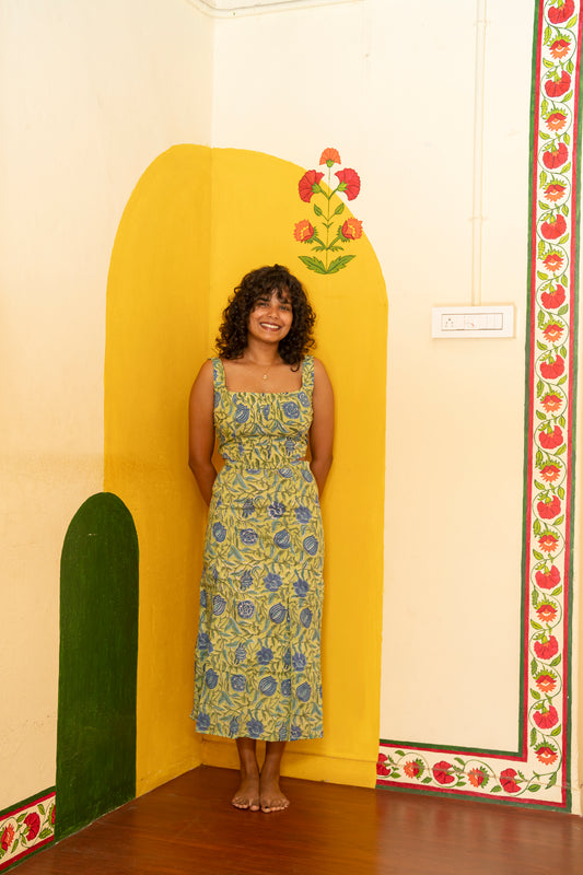 A woman stands barefoot in a room corner wearing a sleeveless yellow and blue floral print maxi dress, posing in front of a painted yellow archway.