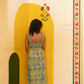 A woman stands barefoot in a room corner wearing a sleeveless yellow and blue floral print maxi dress, posing in front of a painted yellow archway.