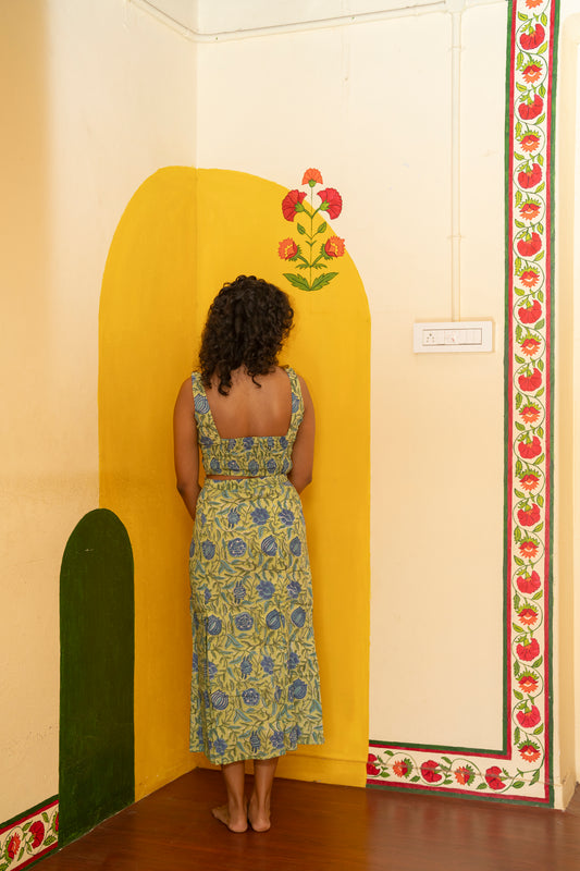 A woman stands barefoot in a room corner wearing a sleeveless yellow and blue floral print maxi dress, posing in front of a painted yellow archway.