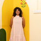 A woman with curly dark hair stands smiling in front of a bright yellow arched backdrop, wearing a sleeveless halter-neck dress with vertical pink and white stripes and a ruffled hem.