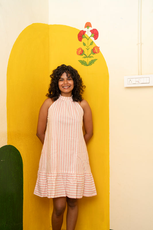 A woman with curly dark hair stands smiling in front of a bright yellow arched backdrop, wearing a sleeveless halter-neck dress with vertical pink and white stripes and a ruffled hem.