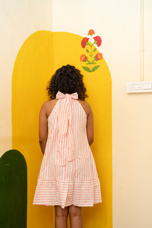 A woman viewed from behind wearing a pink and white striped halter dress with a large bow at the back, standing in front of a bright yellow painted wall with a floral motif.