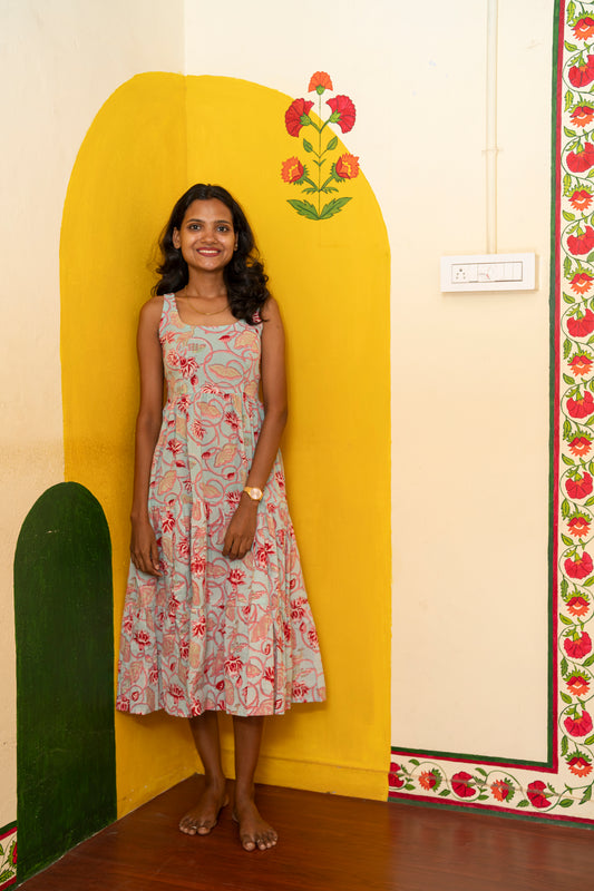 A woman stands wearing a sleeveless,  floral midi dress with thin straps. She is standing in a decorated room with a yellow arched wall, a green arch, and a wooden floor.