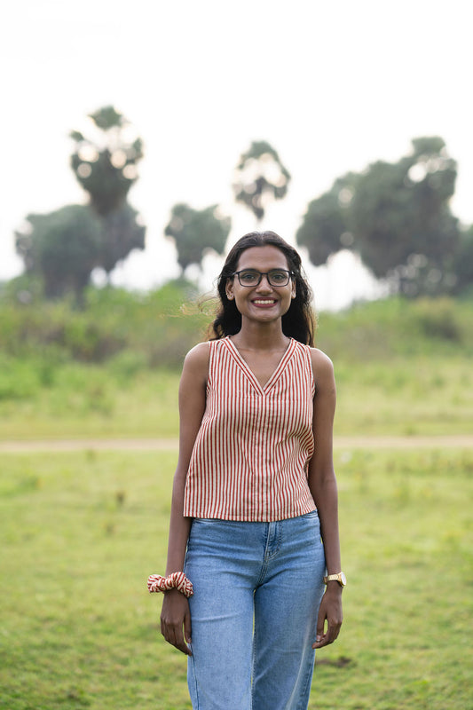 A woman wearing a sleeveless red and white striped top and blue jeans stands outdoors in a grassy field with trees in the background.