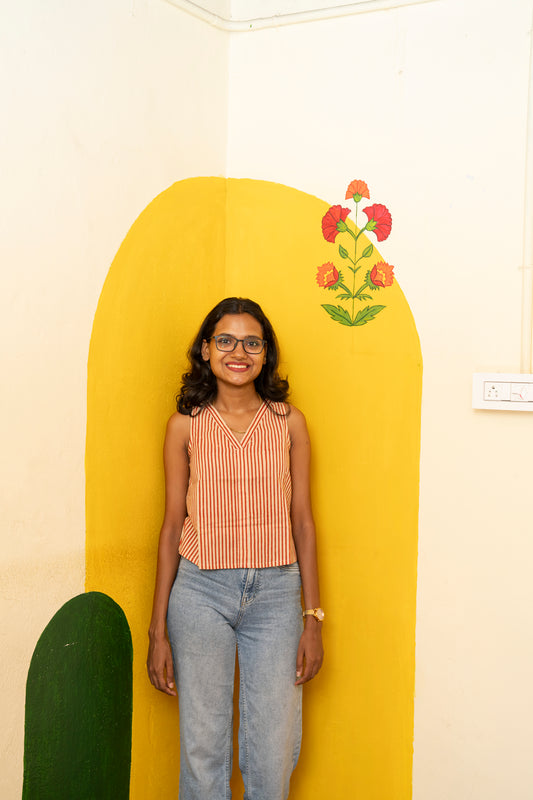 A woman wearing a sleeveless red and white striped top with light blue jeans stands in front of a yellow painted wall with a floral motif.