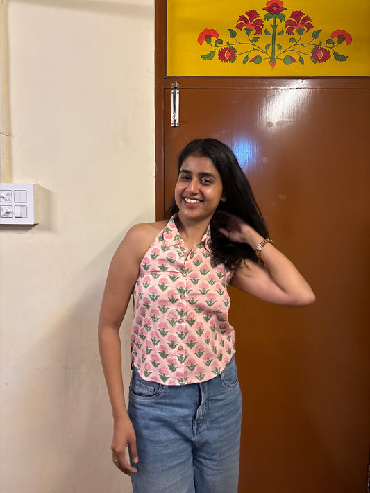 Woman posing in a room with a floral curtain and wooden door wearing pink shirt with floral patterns.