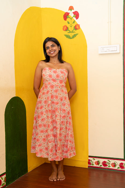 A woman stands barefoot indoors wearing a strapless midi dress with a pink and orange floral print on a light beige background. She is posed in front of a bright yellow painted arch on a cream wall.