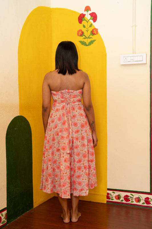 A woman stands with her back to the camera wearing a strapless pink floral midi dress, positioned in front of a yellow painted wall corner with a floral motif.