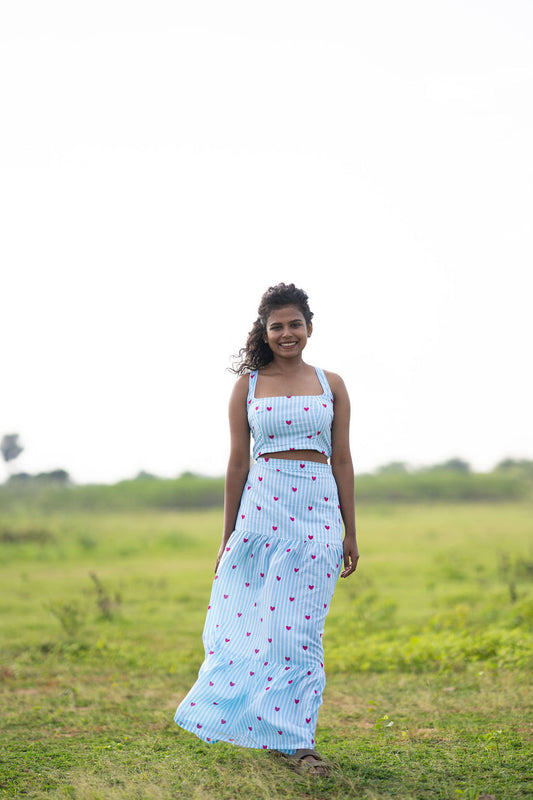 A woman wearing a light blue and white checkered two-piece crop top and maxi skirt set with red polka dots, standing in a grassy field.