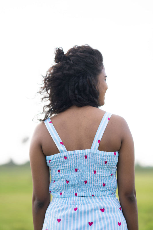 A woman viewed from behind wearing a light blue and white vertically striped dress with pink heart prints, standing in an outdoor grassy field.