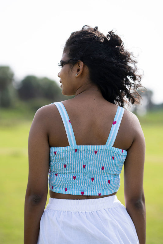 A woman viewed from behind wearing a light blue and white striped crop top with small red heart prints and a white skirt, standing in an outdoor grassy field.