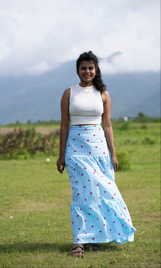 A woman standing in a grassy field with mountains in the background, wearing a white sleeveless crop top and a long light blue maxi skirt with thin vertical stripes and small pink heart prints.