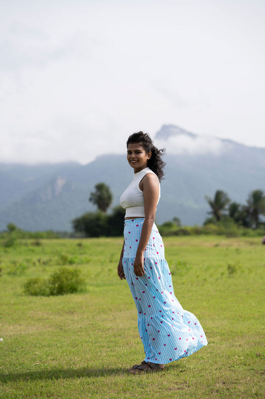 A woman stands in a grassy field with mountains in the background, wearing a white sleeveless crop top and a long light blue skirt with a small red and yellow pattern.