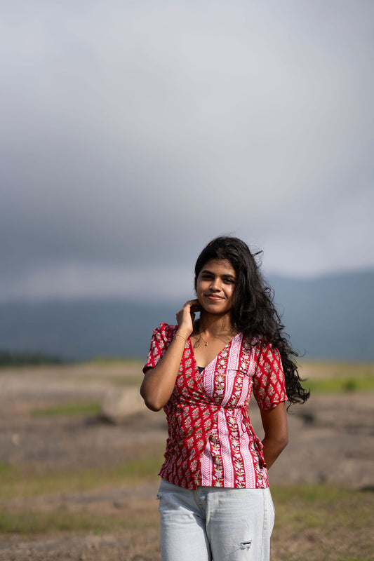 A woman with long dark hair stands outdoors in a dry, grassy landscape under a cloudy sky, wearing a red and white patterned short-sleeve top and light blue jeans.