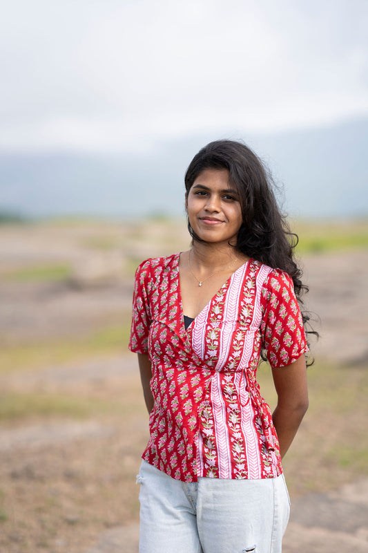 A woman wearing a red and white patterned short-sleeve top with white pants, standing outdoors in a dry, grassy landscape.